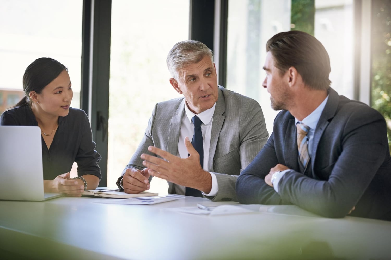 Three professionals sit at a table having a meeting. The central figure gestures while speaking. They appear engaged and focused, conveying collaboration.