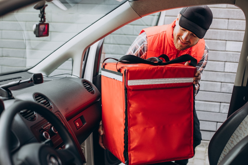 A delivery driver in a red vest smiles while placing a large red insulated bag in a car. The interior and dashboard of the car are visible.