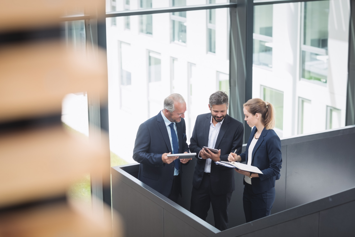 Three professionals in suits stand discussing documents indoors, near large windows. They appear focused and engaged in conversation.