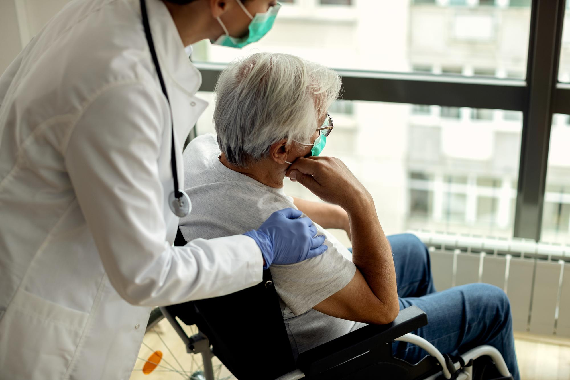 A doctor in a white coat and mask comforts an elderly patient in a wheelchair by a large window. They both wear masks, conveying a sense of care.
