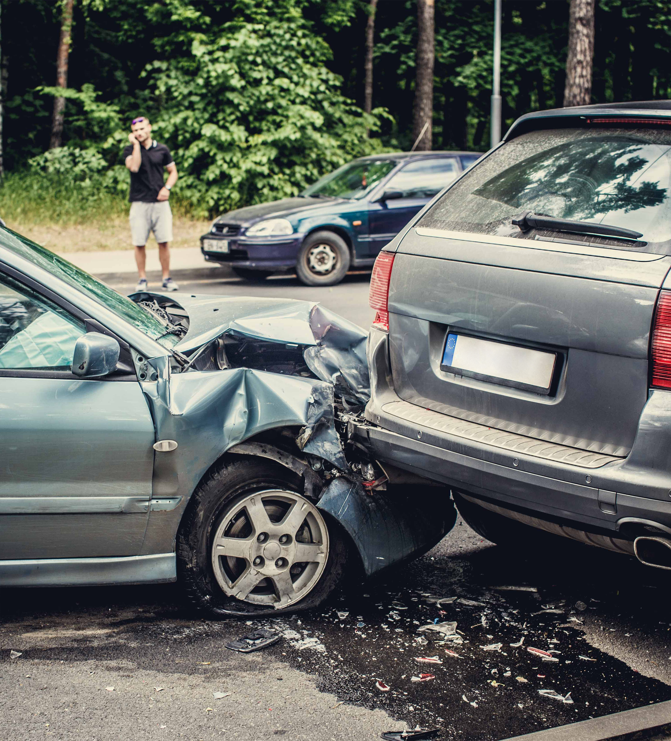 Two cars involved in a rear-end collision on a forested road. The front car is severely damaged. A man in the background makes a phone call, appearing concerned.