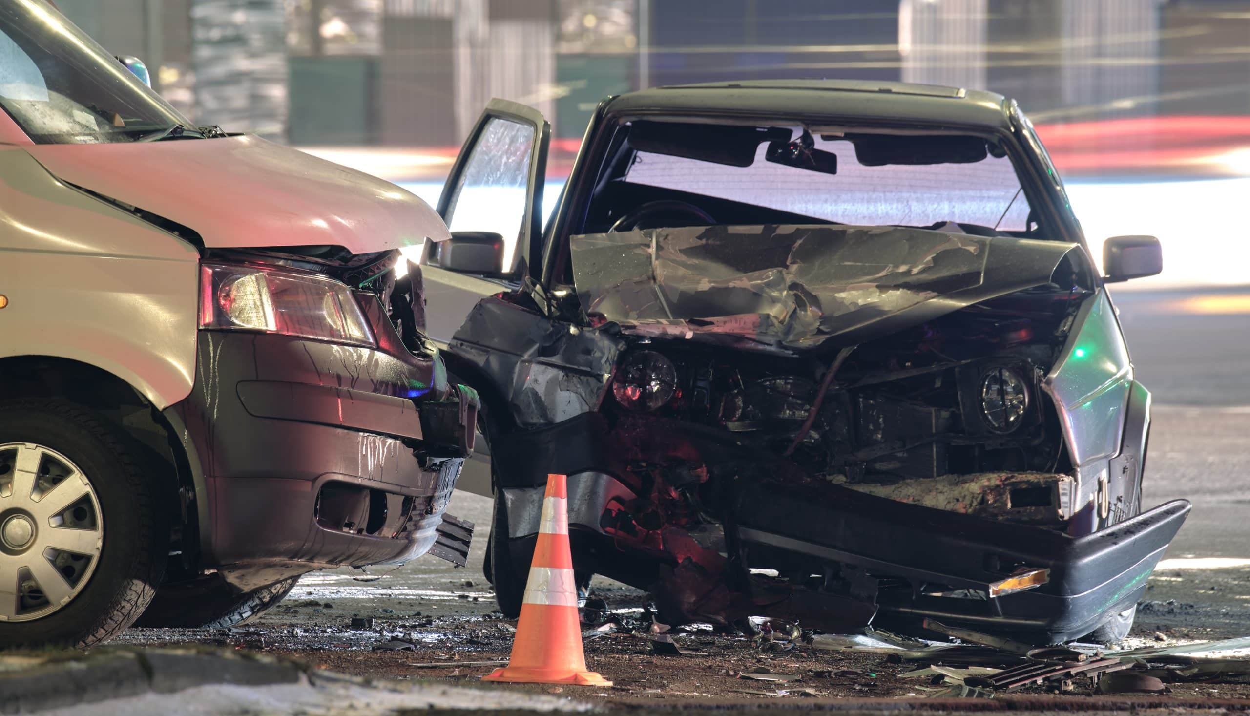 Two heavily damaged vehicles in a nighttime crash. The front ends are crumpled, and debris is scattered. A traffic cone is nearby, adding urgency.