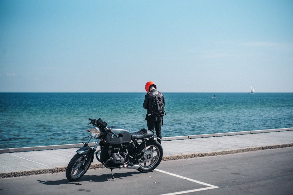 A man stands next to a motorcycle by the ocean, serving as a reminder of the hazards of distracted driving for motorcyclists.