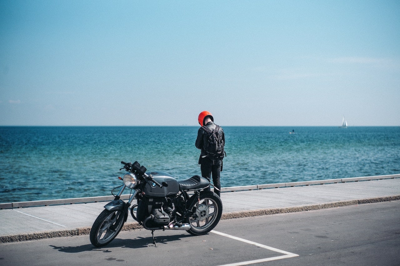 A man stands next to a motorcycle by the ocean, serving as a reminder of the hazards of distracted driving for motorcyclists.