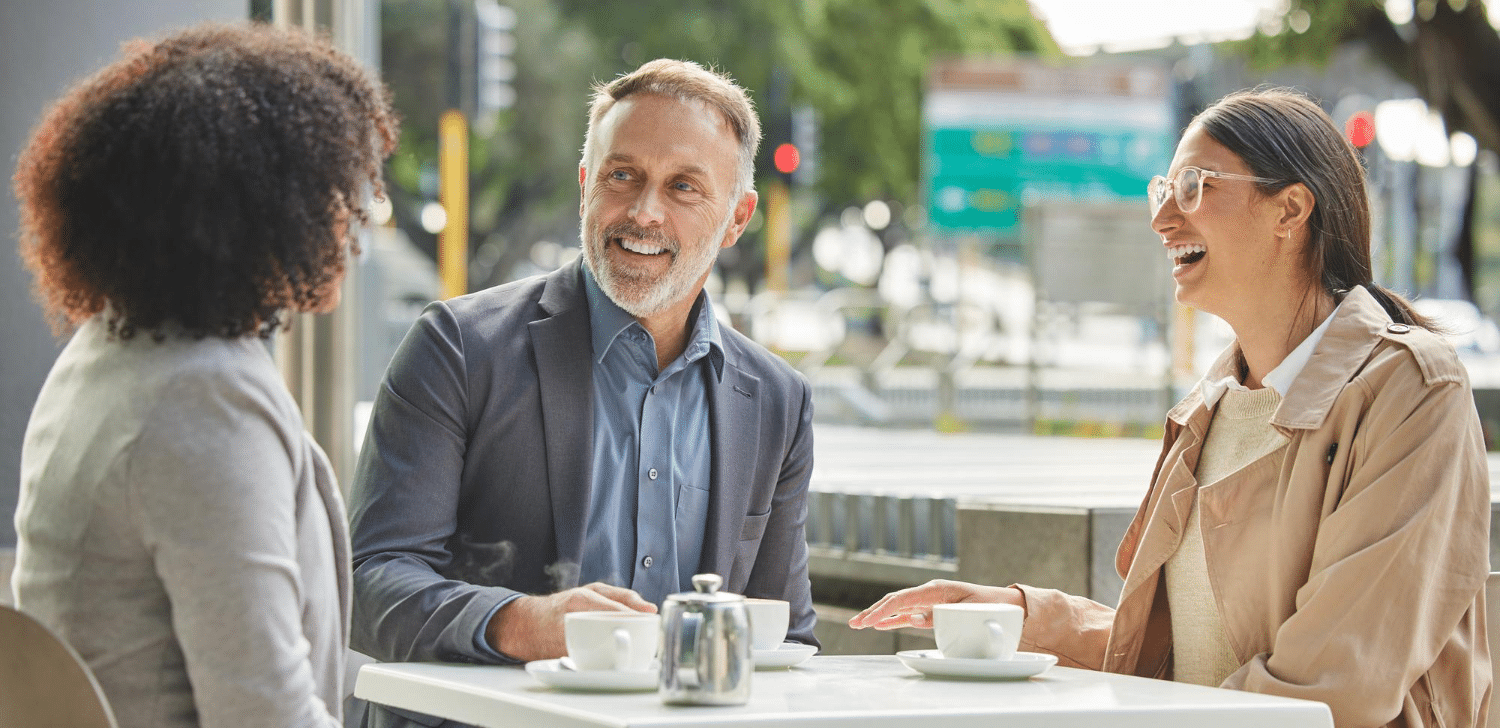 Three people sit at an outdoor café, smiling and engaged in conversation. Cups of coffee and a teapot are on the table. The mood is friendly and relaxed.